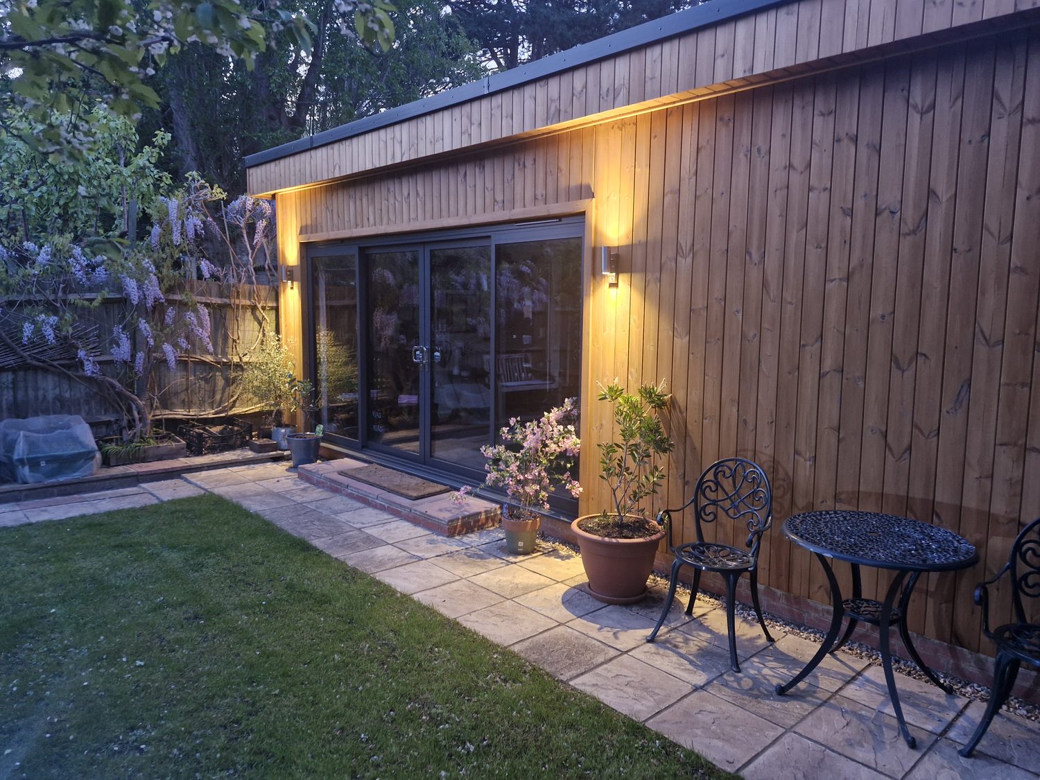 A timber-clad Crown Sandringham garden room lit warmly at dusk, with full-height glazing and wisteria flowering in the garden.