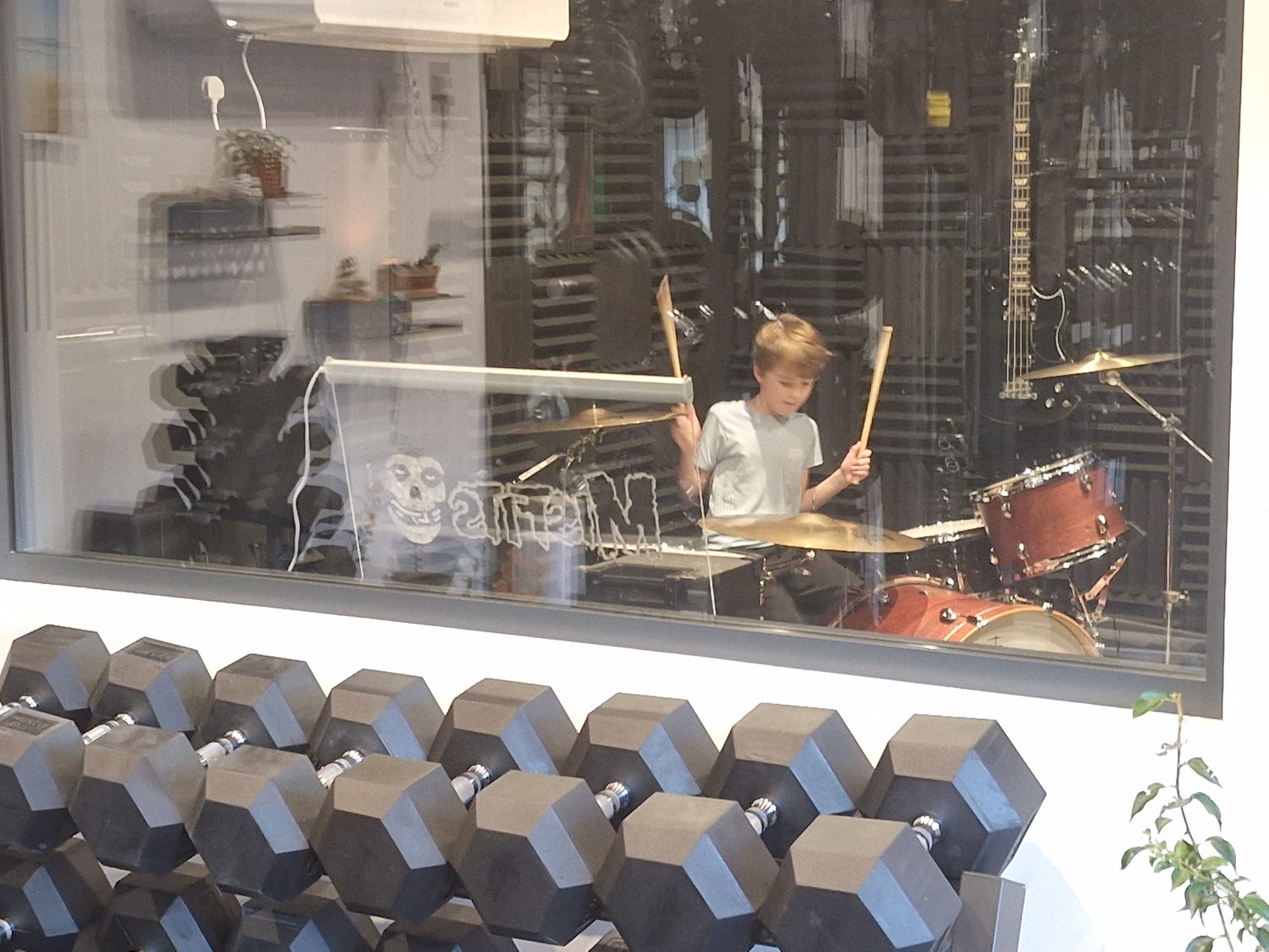 A drum kit in the soundproofed studio, viewed through the gym window past a rack of hex dumbbells.